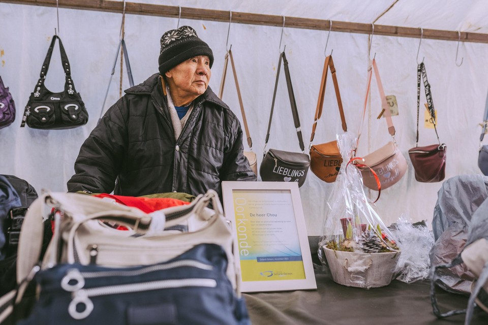 Marktkoopman Sunling Chou achter zijn kraam vol lederwaren. In het midden de gemeentelijke oorkonde die hij zojuist heeft ontvangen van wethouder Leon Geilen van Sittard-Geleen.