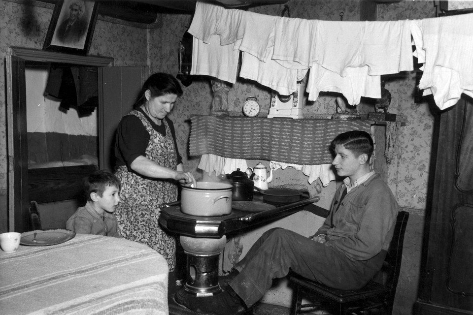 Eenkamerwoning voor een vrouw en vijf kinderen, in 1950. Terwijl de was droogt, roert moeder in een pan op een plattebuiskachel. 